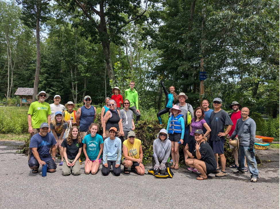 A group of smiling, muddy, people pose with a very large heap of green and brown vegetation.