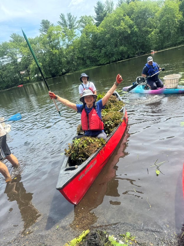 In the foreground, a smiling woman in a red kayak with several baskets full of water chestnuts. In the background, a few other kayers on the pond with their baskets.