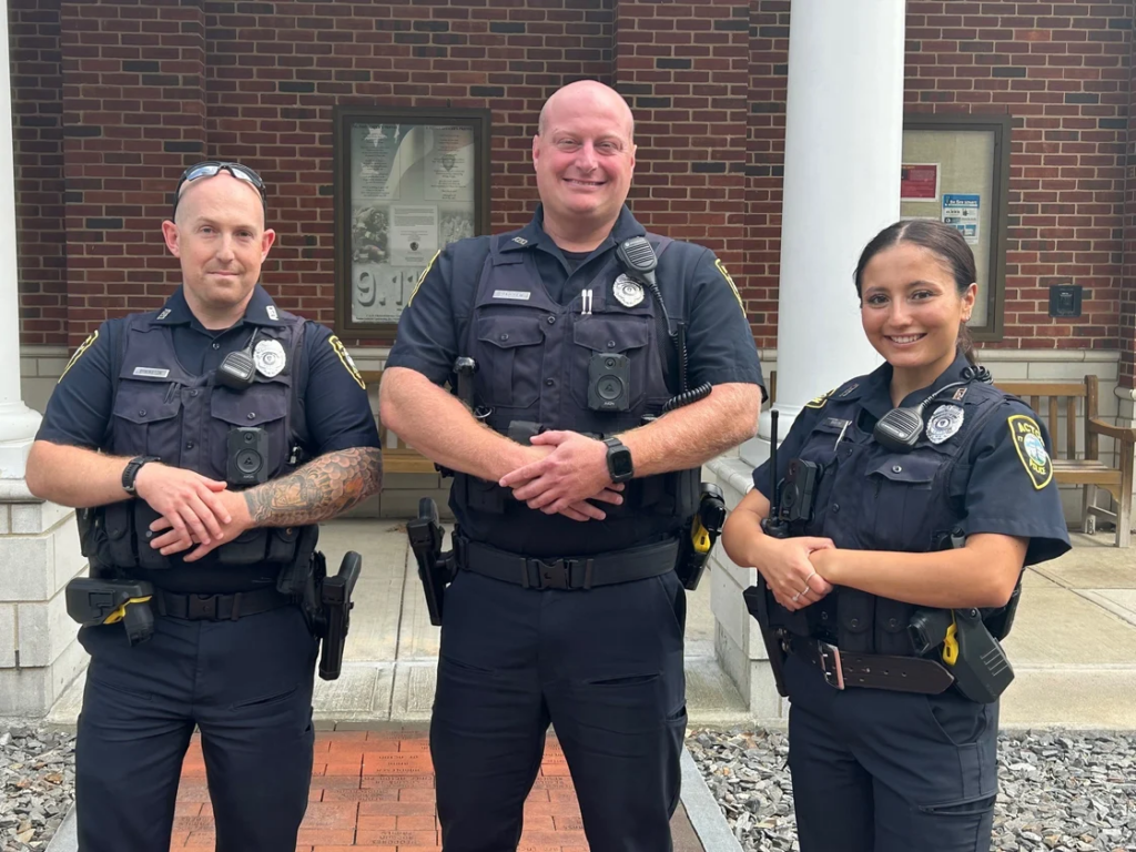 Three uniformed police officers stand in front of the Acton Public Safety building.