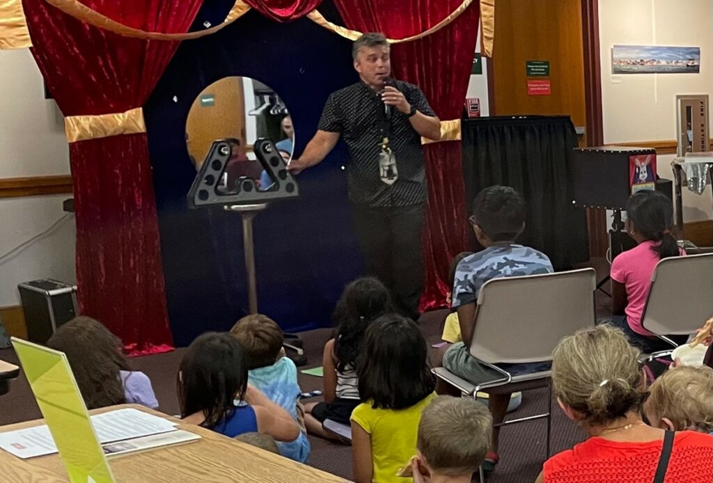 A man on a small stage with a mirror on a stand. Thee are children sitting on the floor and in chairs watching him perform. There is a red velvet curtain in the background and the man speaks into a mic.