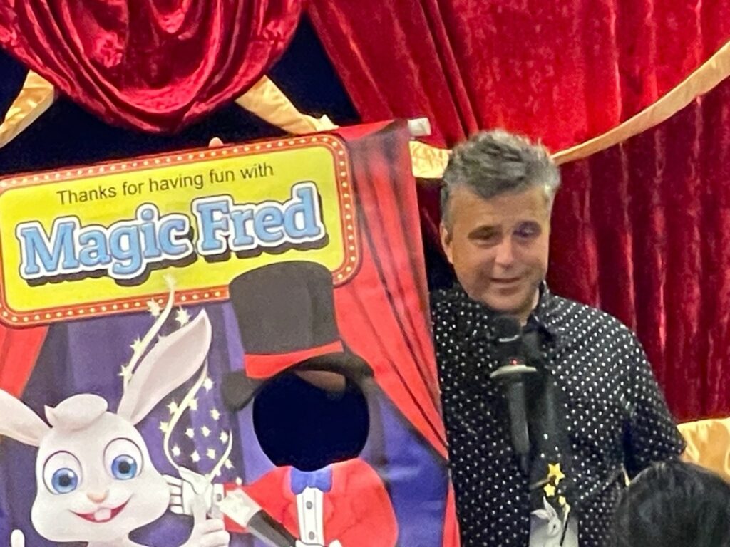 A man stands on the stage with a poster that says "Thanks for having fun with Magic Fred." The poster shows a cartoon bunny and a cutout for a face.