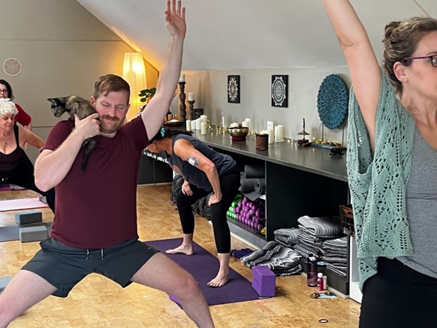 A man in shorts and red t-short does a yoga pose with one hand up, and his other hand holding a kitten on his shoulder.