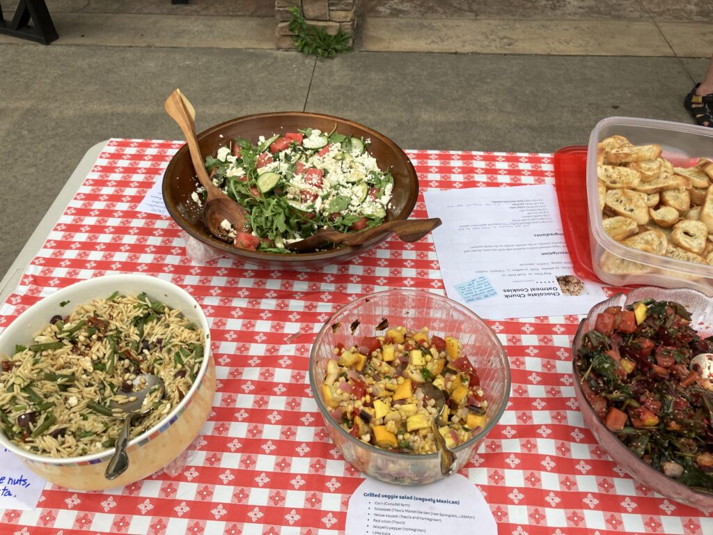 A red-checked tablecloth with several salads. Each salad has a paper copy of the recipe under its bowl.