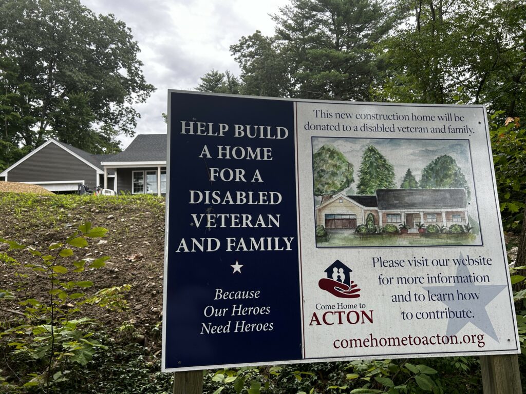 A sign for Come Home to Acton in front of a house under construction. The sign says: Help build a home for a disabled veteran and family. Because Our Heroes need Heroes. This new construction home will be donated to a disabled veteran and family. Please visit our website for more information and to learn how to contribute. comehometoacton.org