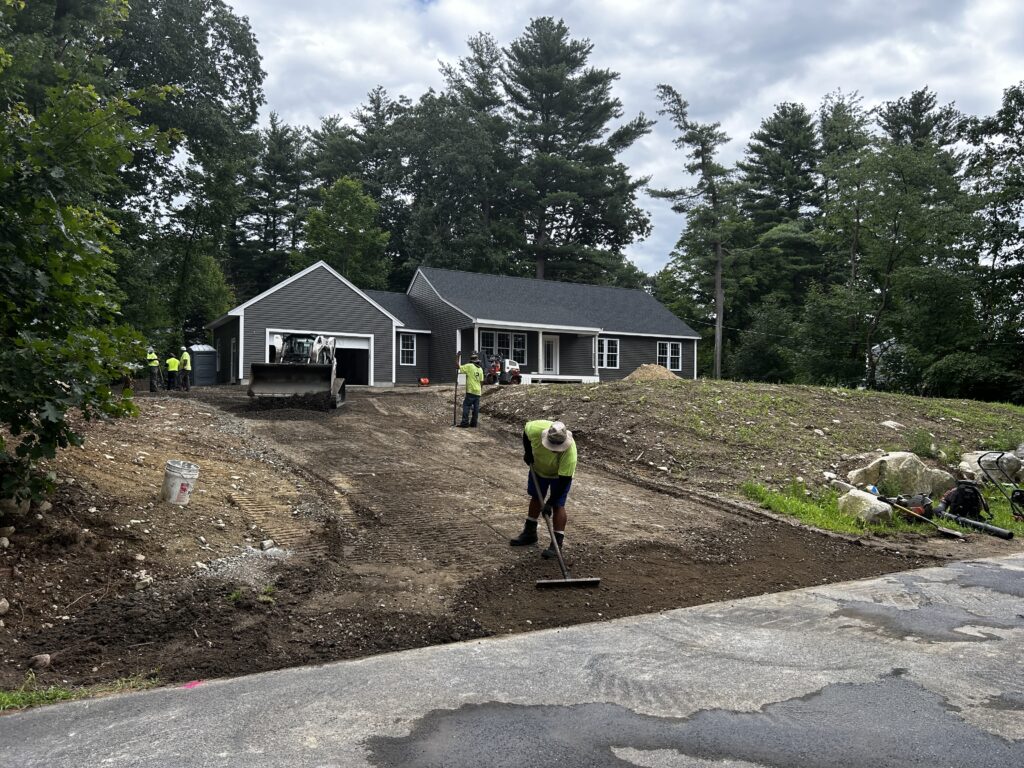 Several workers raking out a dirt driveway. There's also a front end loader in front of the garage.