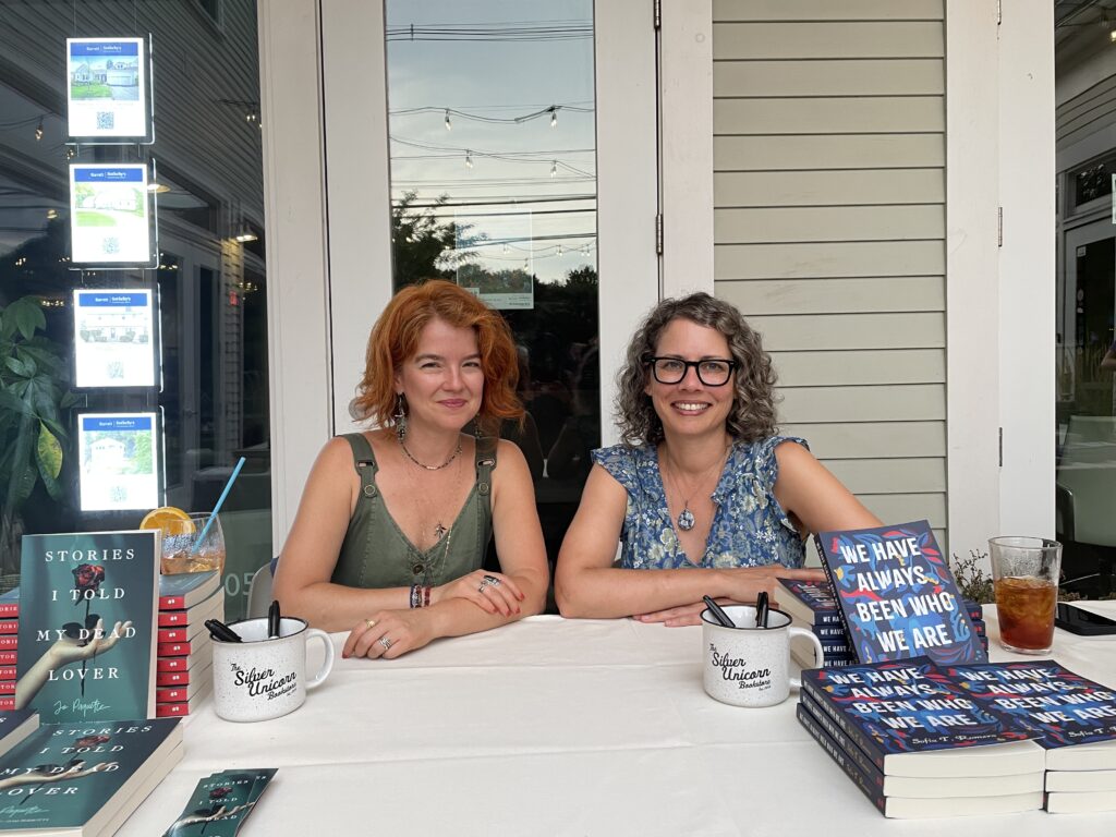 Two smiling women sit at a table. Each has a stack of their newly published books.