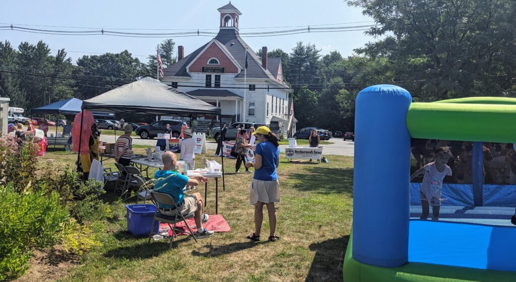 In the foreground, the bouncy house (with bouncing child) on the right, and some peole providing information under canopies. The Boxborough Town Hall is in the background.