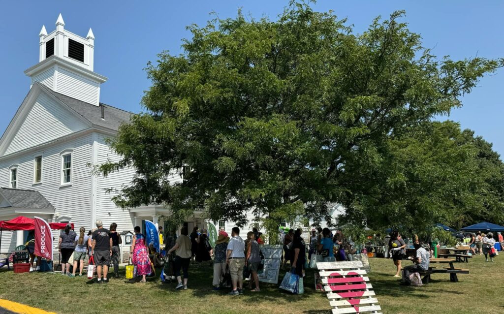 People standing in line in front of the UCC church. Many are standing under a large, shady, tree.