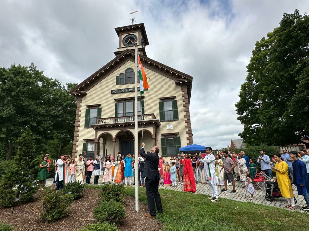 A tall man in a suit raises the Indian flag in front of town hall. Many people are watching.