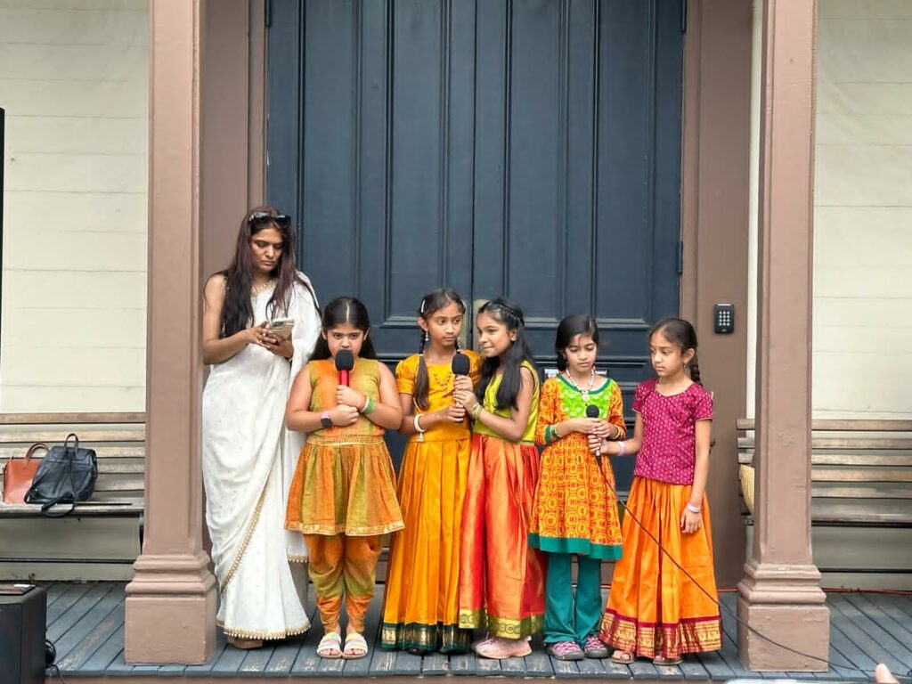 A woman wearing a white sari stands with 5 young girls, all wearing orange Indian clothing. It's rather adorable.