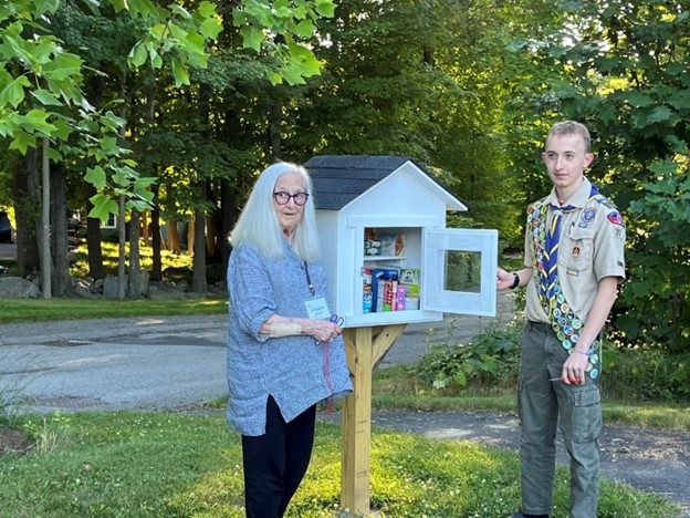 A woman with long grey hair and glasses and a young man in a Boy Scout uniform (with many patches!) stand in front of the new food pantry box. The box is white, has a shingled roof, and stands on a sturdy pedestal. A glass fronted door keeps the weather out, but allows people to see what's available.