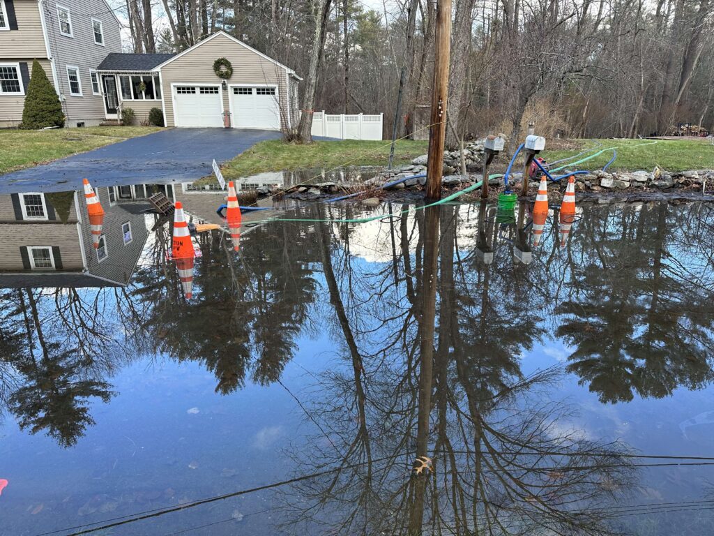 A brown house with a puddle across the road in front ofthe house. The puddle is large enough (and deep enough) to mirror the house and a tree on the property.