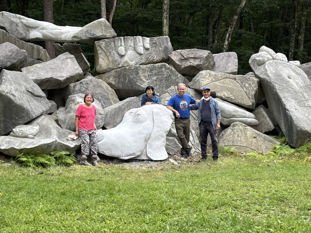 Four adults stand in front of a large pile of boulders, some of which are carved into art. They are standing next to a very large carved dog's face.