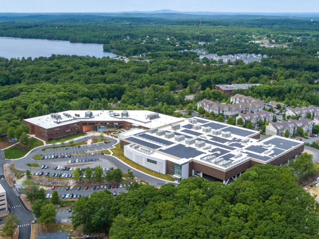 An aerial view of a large cluster of buildings with Nagog Pond in the background.