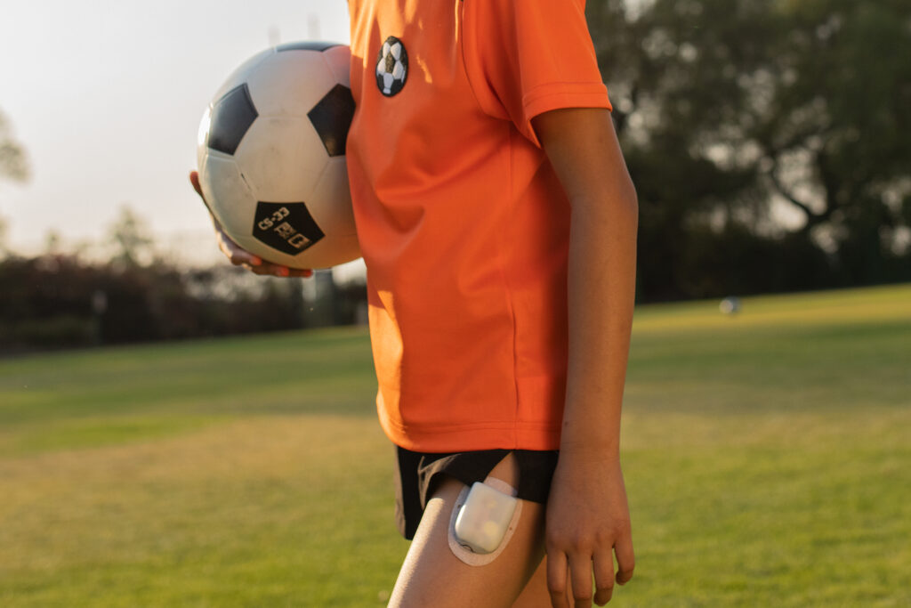 A person, from shoulder to mid-thigh carrying a soccer ball and wearing an orange jersey with an Omnipod attached to their upper thigh.