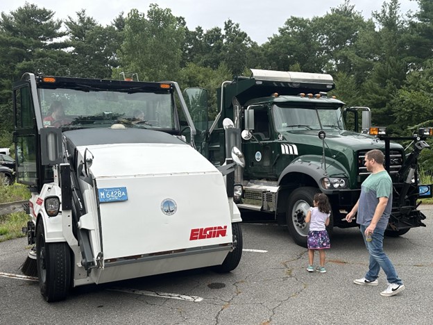 A dad and daughter take a look at a couple of pieces of town equipment. The street sweeper is white and there is someone sitting in the cab (whcih towers above the girl). There's a green dump truck in the background.