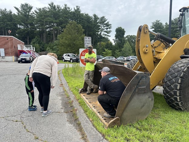 Two men are inside the bucket of a front loader. A mom and child are taking a careful look.