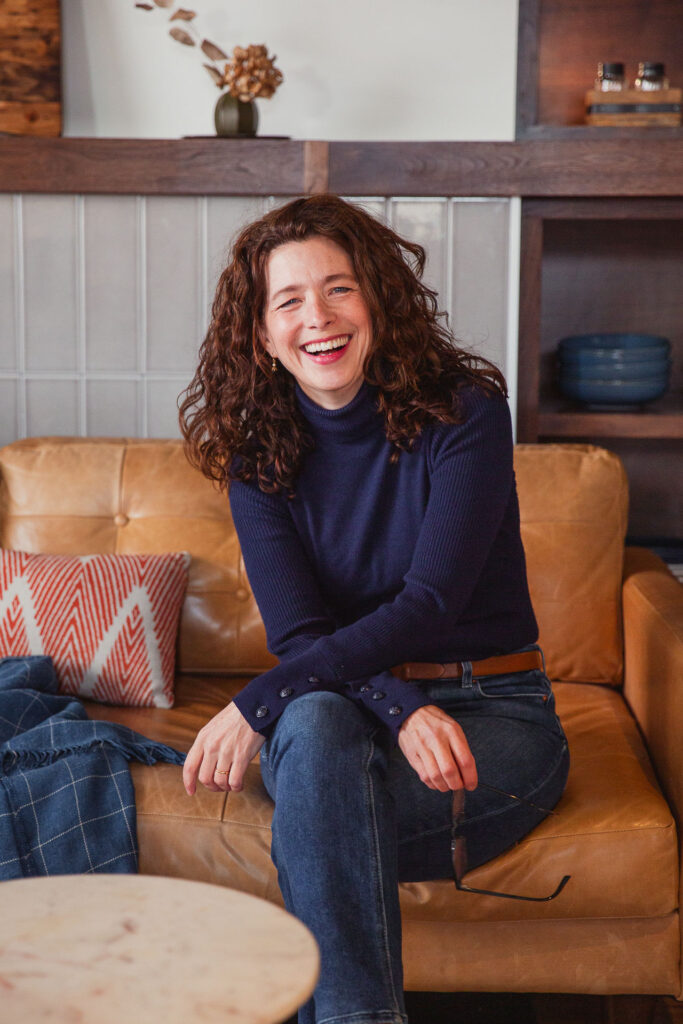 A smiling woman with curly shoulder=length hair and a navy turtleneck poses casually on a leather couch.