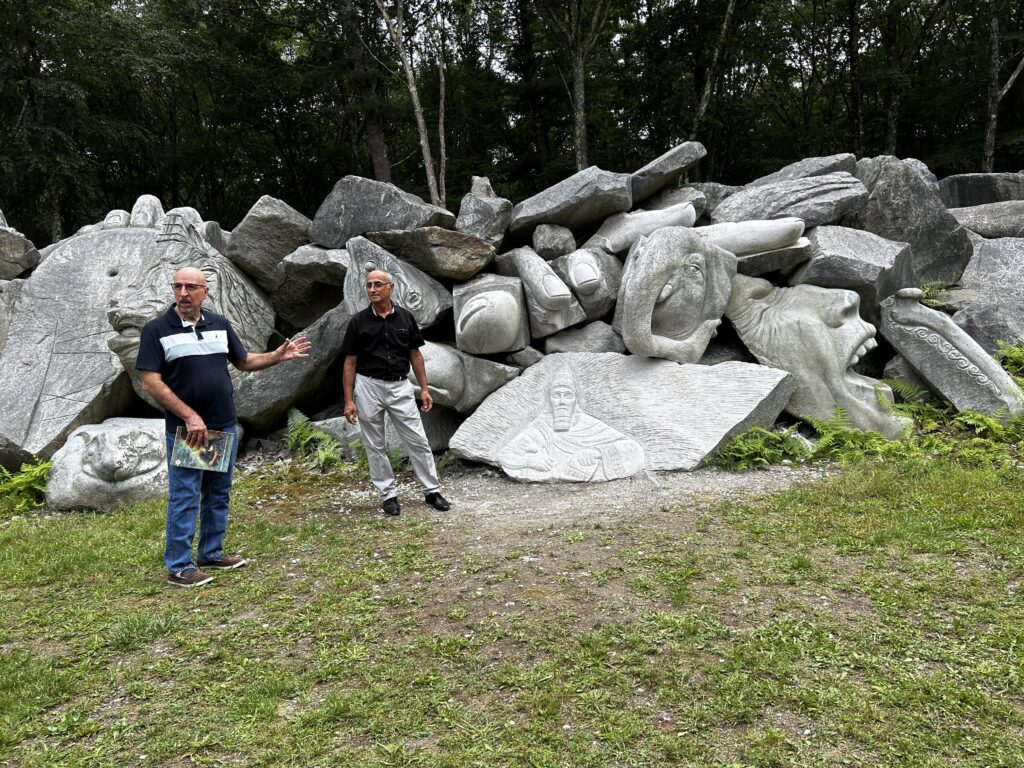 Two men stand before a stone carved with the head of a seated king with a beard and triangular hat. Other nearby boulders are sculpted into an elephant and an open mouthed toothy smile.