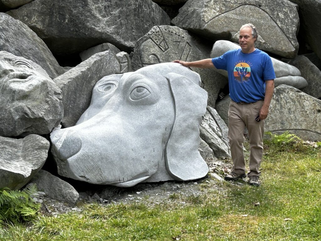 A man with grey hair, beige pants, and a blue t-shirt rests his hand on the top of a large granite sculpture of a dog head, with other granite boulders behind.