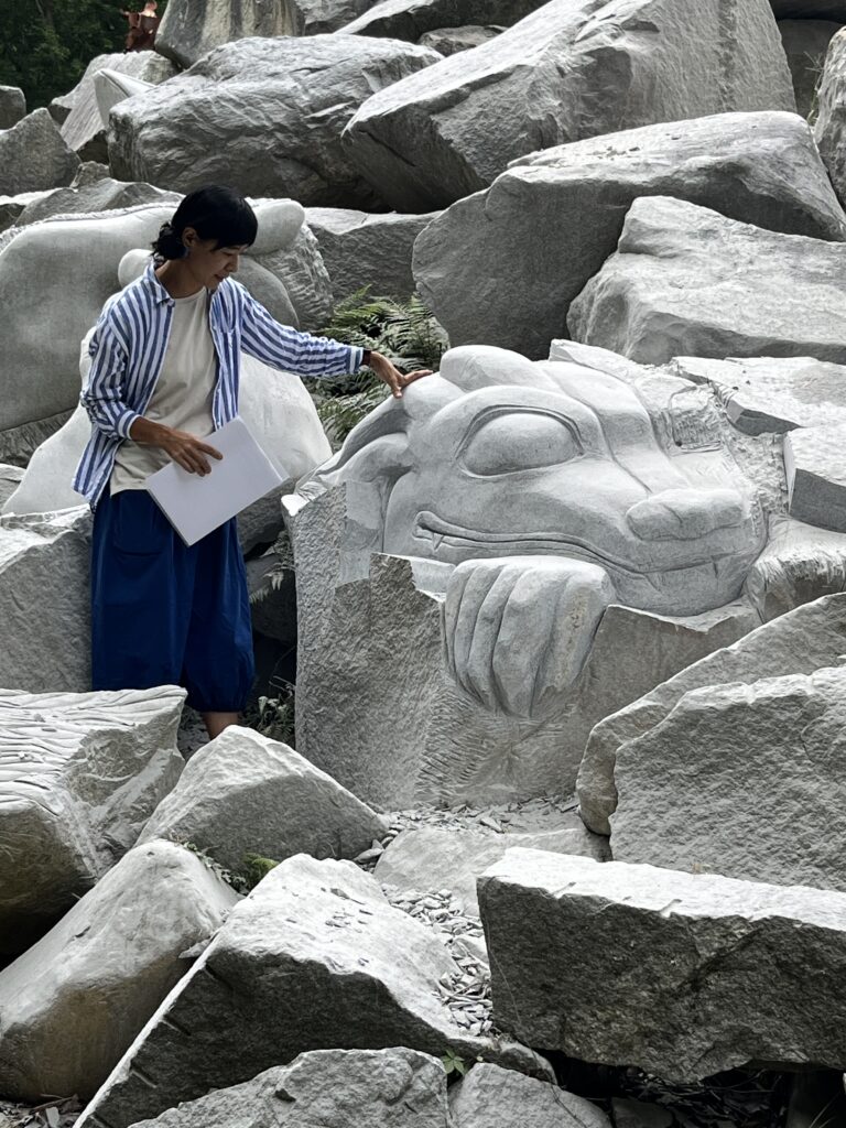 A woman in a blue striped shirt and blue skirt looks toward a large granite head of a baby dragon with a little tooth sticking out of its mouth and a large claw. Other boulders surround the dragon head.