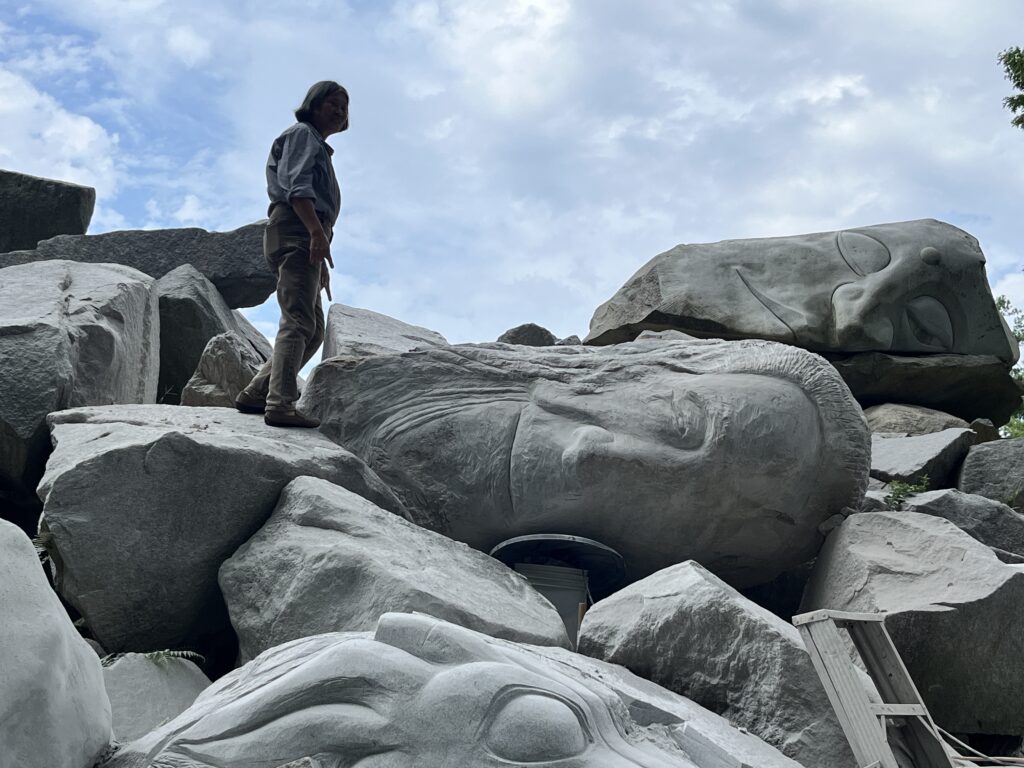 A woman stands silhouetted against blue sky, near three large heads made from boulders, among other boulders in a pile.