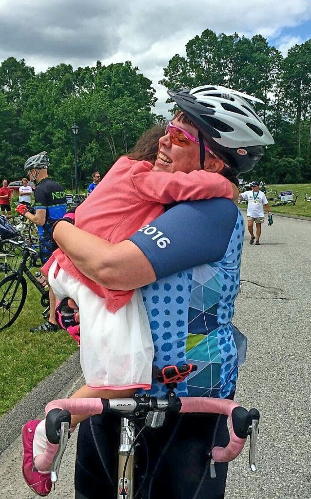 A smiling woman with short brown hair wearing a helmet and cycling gear hugs a child wearing a pink sweater and gauzy skirt. They are leaning against a bike with pink handlebars.