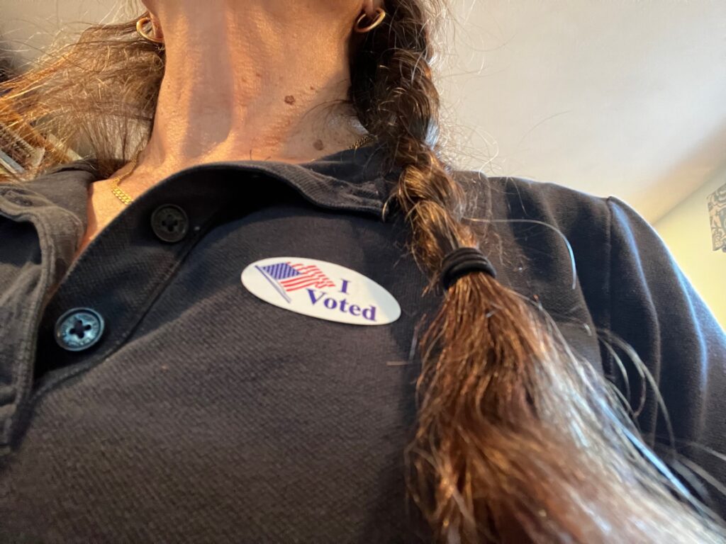 A woman's torso with a grey shirt, a long braid, and an I Voted sticker.