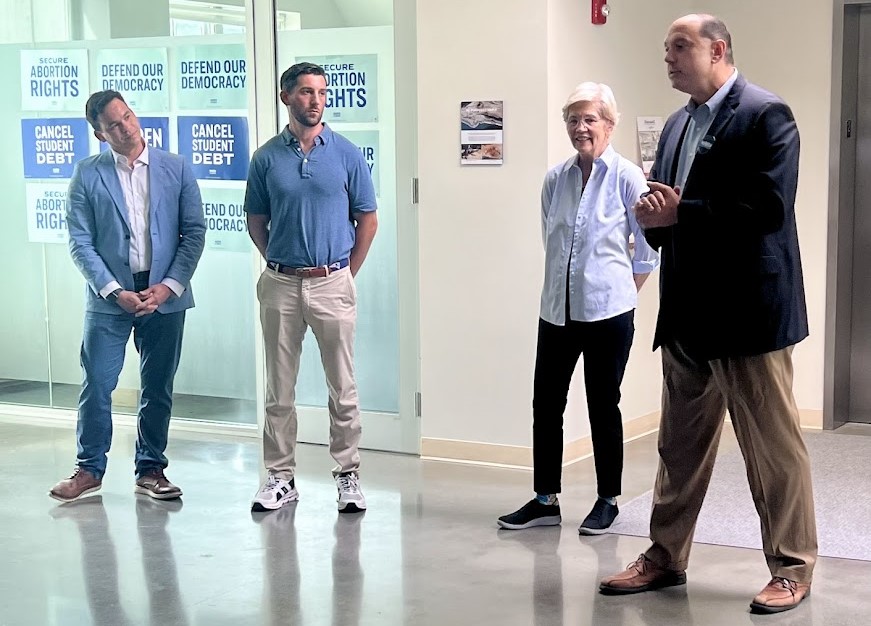 Two casually dressed men and a woman listen to a tall man wearing a suit jacket.