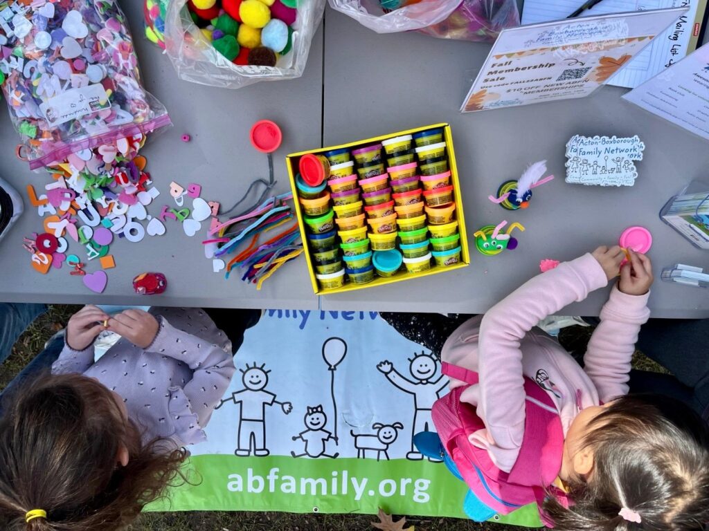 The tops of the heads of two young children standing in front of a table filled with crafting supplies such as paper letters, pompoms, pipe cleaners, and tiny Play-Doh containers.