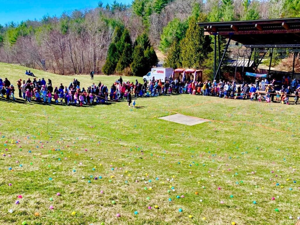 A crowd of children and their adults are lined up at the bottom of the NARA Park amphitheater. In the foregroud, the lawn on the hill is covered with brightly colored egs.