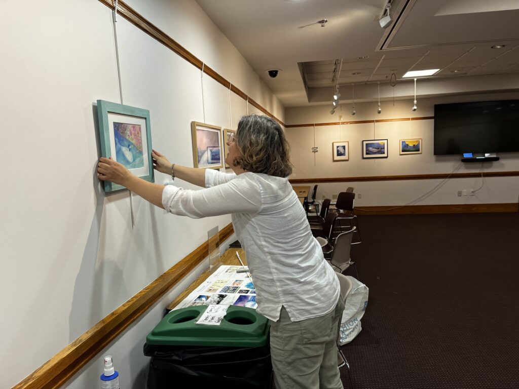 A woman with shoulder-length graying hair hangs a picture on the wall of the Acton Memorial Library meeting room. The picture is a drawing of a white whale swimming in the sea.