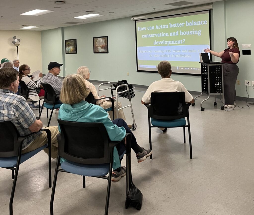 A woman with long brown hair and bangs speaks at a podium in front of an audience at the Senior Center. She stands next to a screen that asks "How can Acton better balance conservation and housing development."