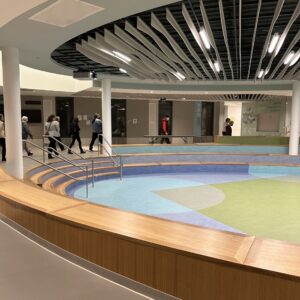 People walking around a round sunken area. There is a low wooden wall and a few steps leading down to the lower area, which has a design of blue and green circles and squares.