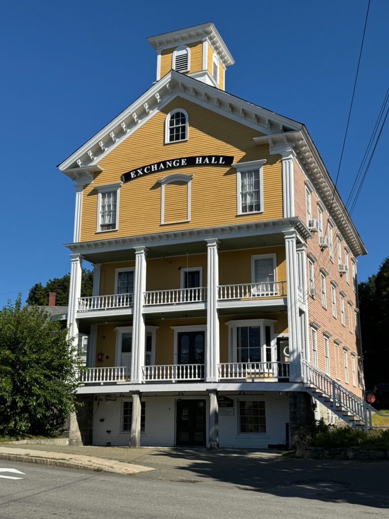 A very large three story building, painted yellow and white. The bottom two stories have white wrap-around balconies and there is cupola on top. A large Exchange Hall sign is near the top of the building.