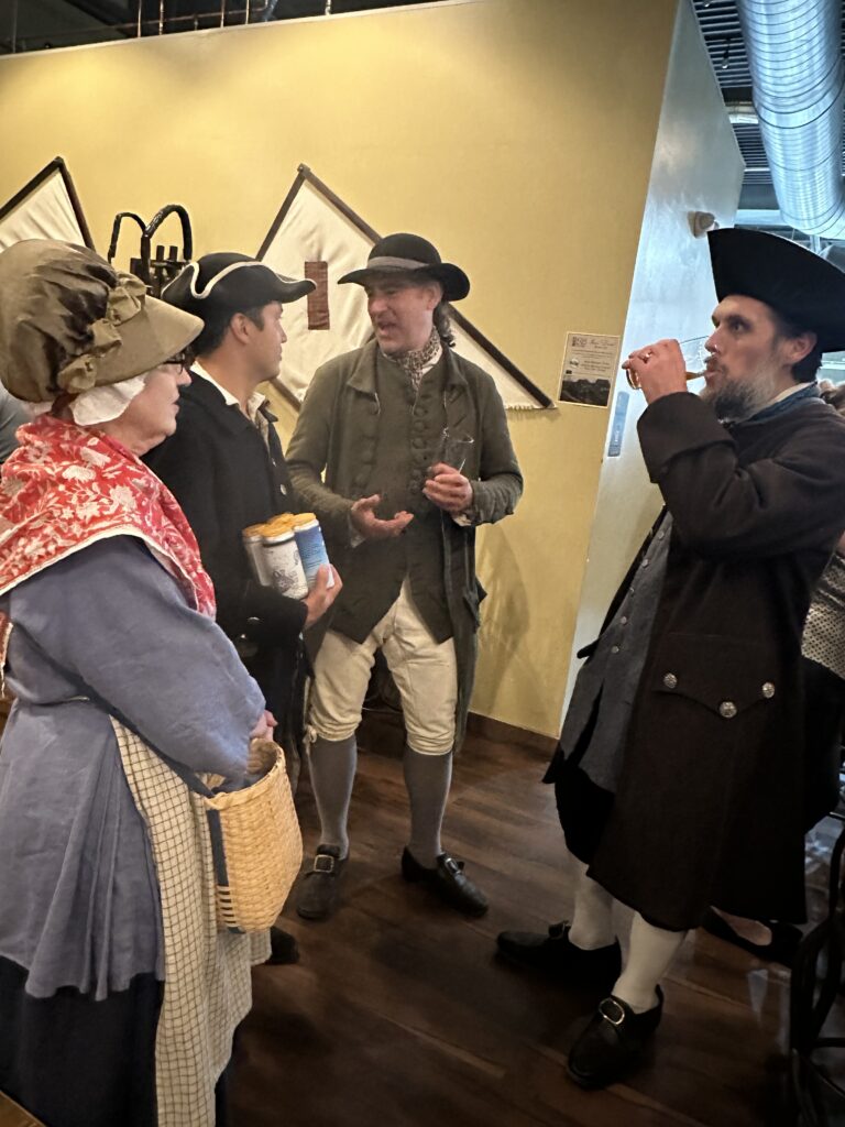 A group of men and women wearing Colonial garb chat while drinking beer.