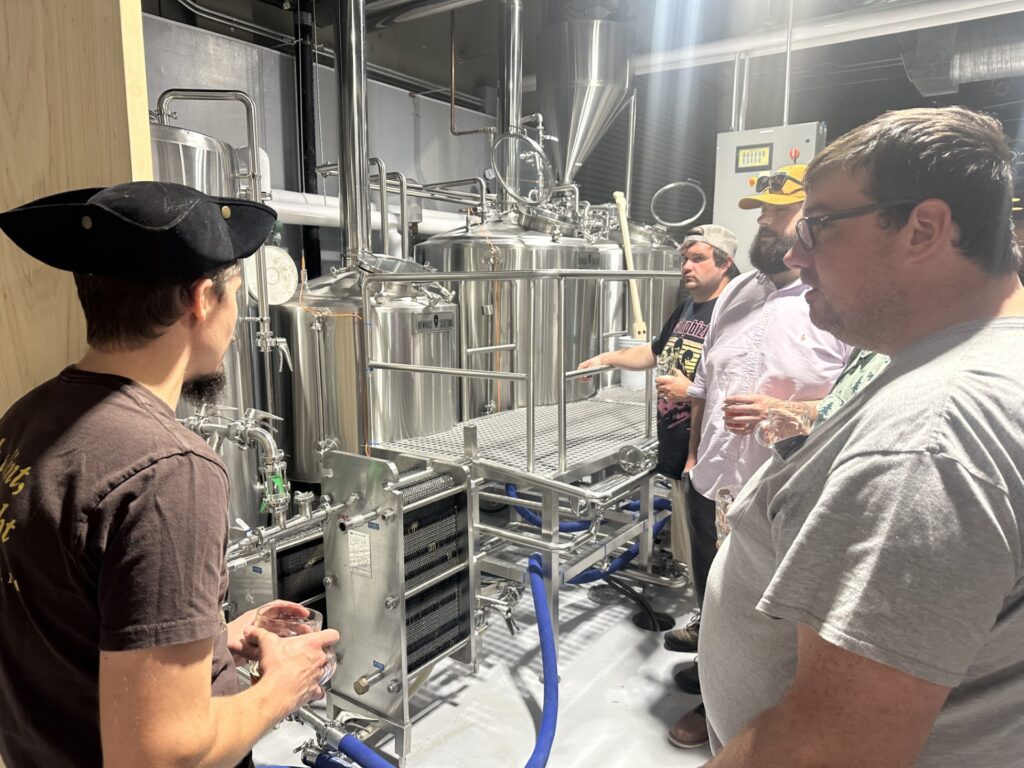 A man wearing a tricorner hat talks to a number of other men. They are standing in front of a steel vats and tubes (beer making equipment, no doubt).