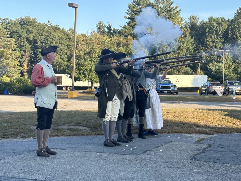 A group of four men and one women in Colonial dress have just fired muskets -- there's smoke rising from the old-fashioned guns. A fifth man, also in Colonial clothing stands by to give instructions.