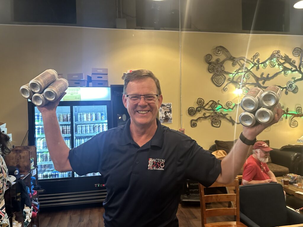 A smiling man wearing an Acton 250 golf shirt hefts a 4-pack of Isaac Davis Brown Ale in each hand.