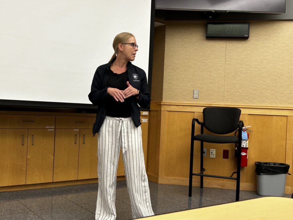 A tall blonde woman, with a black jacket (that has a Clinician patch) and black and white striped pants is speaking in the front of a room.