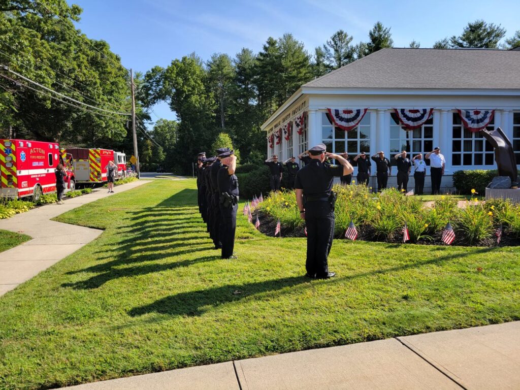 Many of Acton's first responders line up in a salute in front of the Acton Public Safety building. There are two bright red EMT vehicles in the background.