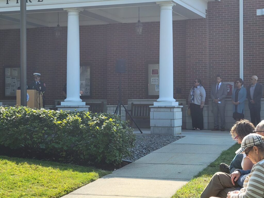 The fire chief, in her full uniform, is speaking at a podium under the eaves of the Public Safety Building. There are several other people off to the right and seated in the audience.