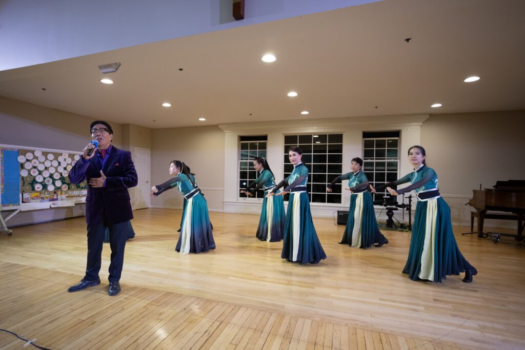 A group of women wearing long teal and white gowns dances in a wood-floored hall. A man sings in the foreground