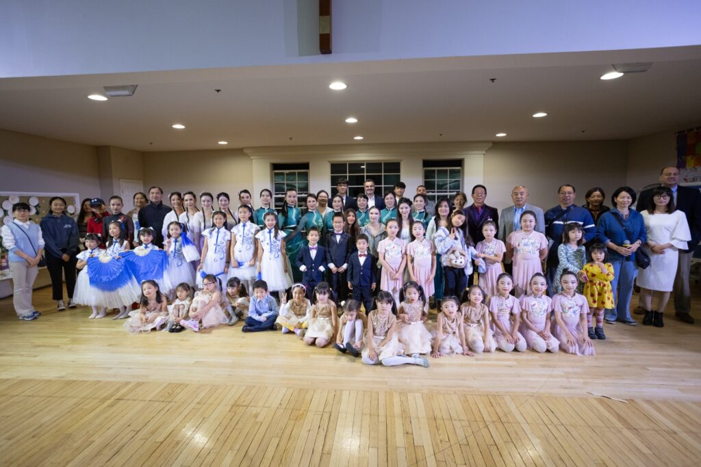 Multiple groupls of performers sit or stand in the performance space. The groups include groups of children and adults in dance outfits, and speakers wearing suits or other street clothing.