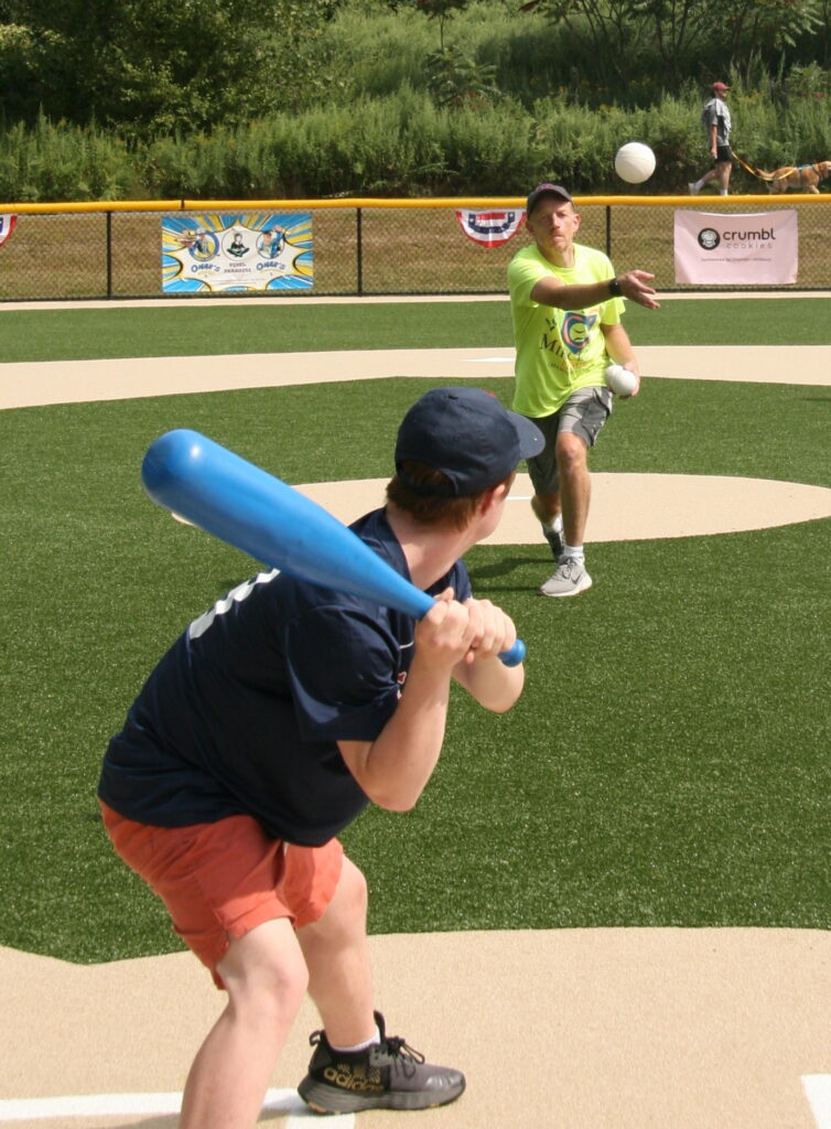 A man, wearing a Miracle League t-shirt, pitches to a player who is holding a bright blue bat.