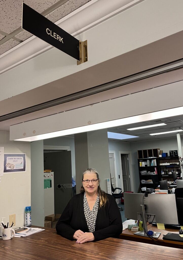 A woman stands in front of the Acton Town Clerks office. You can see the crowded office in the background.