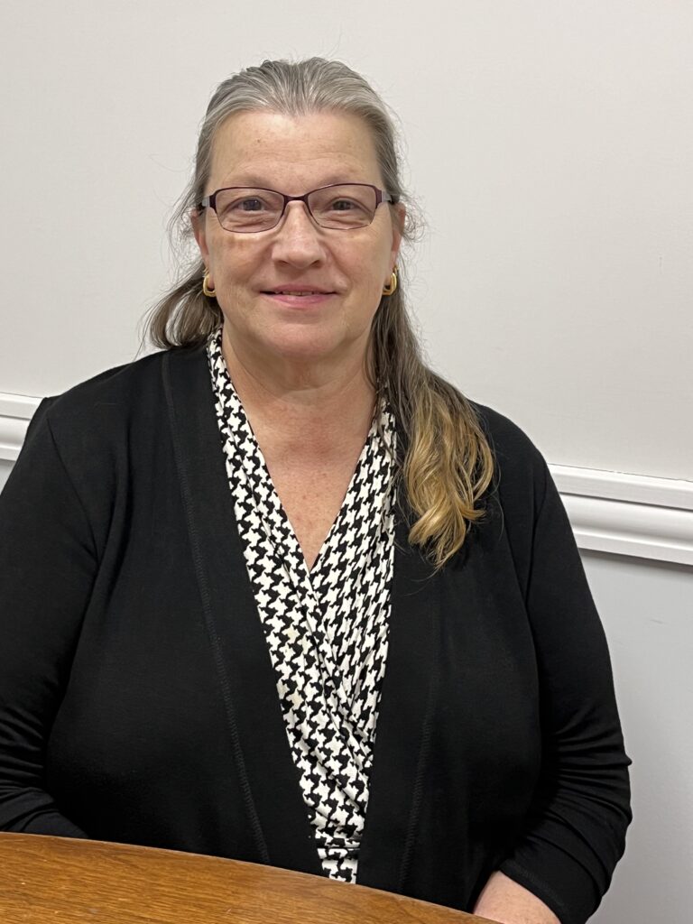 A woman with graying hair and glasses sits at a table. She is wearing a black jacket and black and white houndstooth-checked shirt.