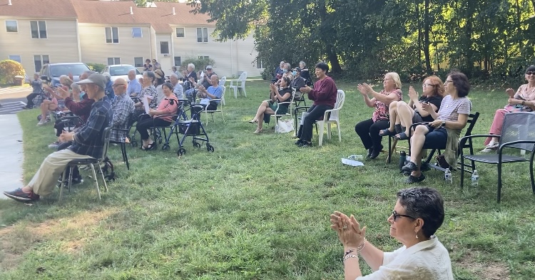 A group of people clapping. People are sitting in lawn chairs on the grass.