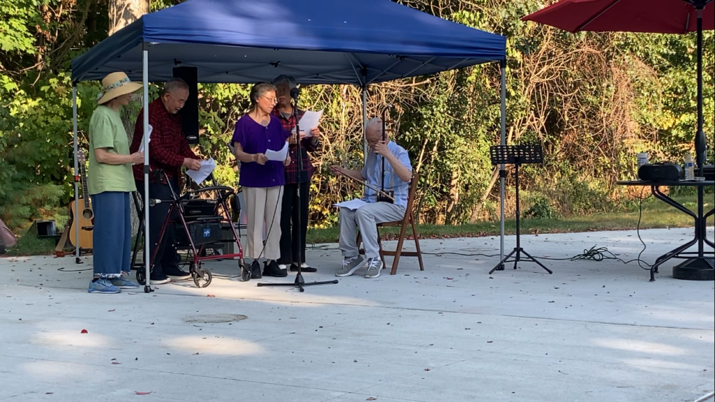 Four older Chinese people stand under a small tent. A man in a blue shirt is seated off to the side playing a T-shaped instrument (with the shorter section in his lap) with a bow.