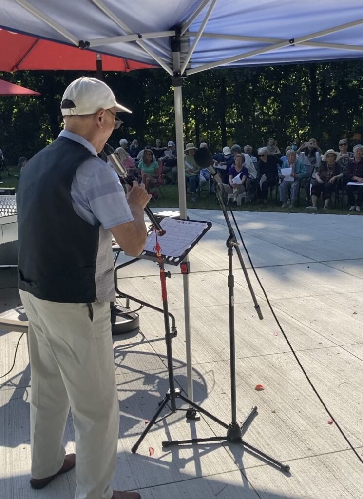 A man, from the back, wearing a baseball cap and a vest plays a black flute. The audience, in lawn chairs, is in the background.